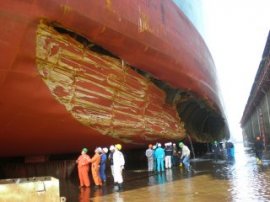 People observe a big tanker with a giant gash with its hull in dry-dock.